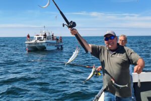 Man staat op boot met hengel in zjin hand. Aan de lijn zitten vissen, die de man trots laat zien.