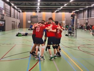 Volleybalteam in rode shirts viert samen een punt in een sporthal, spelers staan in een kring en omhelzen elkaar.